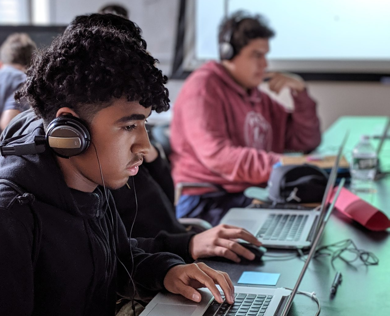 A student sitting at a laptop with headphones around his ears, surrounded by other students deep in thought. He is black, with short, curly hair, wearing a black hoodie.